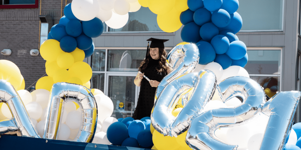 student standing underneath balloon arch with graduation cap